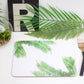 A placemat with a palm leaf design, laid out on a table with a green plant and a wooden cutlery set on top of it.