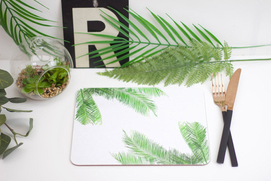 A placemat with a palm leaf design, laid out on a table with a green plant and a wooden cutlery set on top of it.