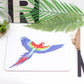 A colorful parrot placemat with a design, placed on a table with a green plant, a coaster, and a pen.
