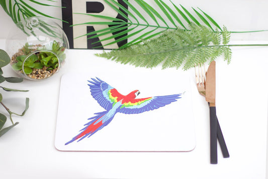 A colorful parrot placemat with a design, placed on a table with a green plant, a coaster, and a pen.