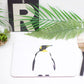A white placemat with a penguin illustration, flanked by cutlery and surrounded by decorative foliage.