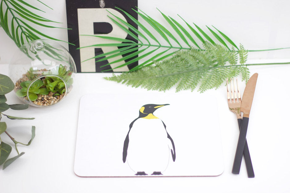 A white placemat with a penguin illustration, flanked by cutlery and surrounded by decorative foliage.