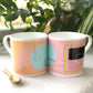 Two bone china mugs with abstract pink and white geometric patterns, one mug is pink with a geometric shape on it, and the other is white with a geometric shape on it, placed on a surface with a golden spoon beside them.