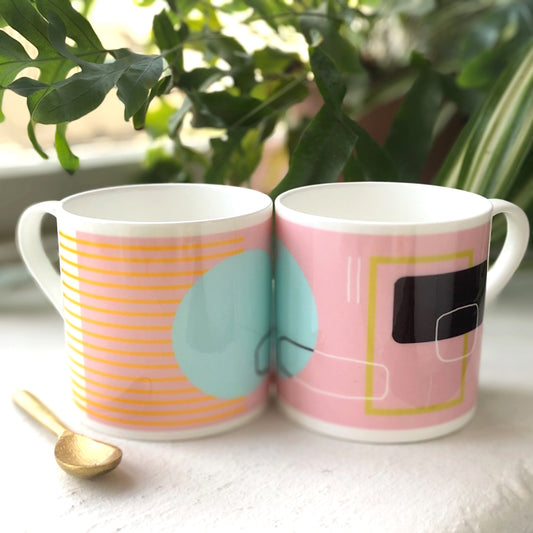 Two bone china mugs with abstract pink and white geometric patterns, one mug is pink with a geometric shape on it, and the other is white with a geometric shape on it, placed on a surface with a golden spoon beside them.