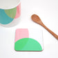 A coaster with a pink and green geometric pattern, a bone china mug with a geometric pattern, and a wooden spoon on a white background.