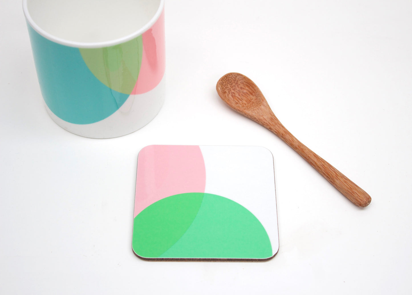 A coaster with a pink and green geometric pattern, a bone china mug with a geometric pattern, and a wooden spoon on a white background.