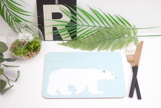 A light blue placemat with a polar bear design, displayed on a table with cutlery and a plant in the background.