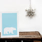 A greetings card with a blue background featuring a silhouette of a polar bear, placed on a wooden surface with a decorative Christmas tree in the background.