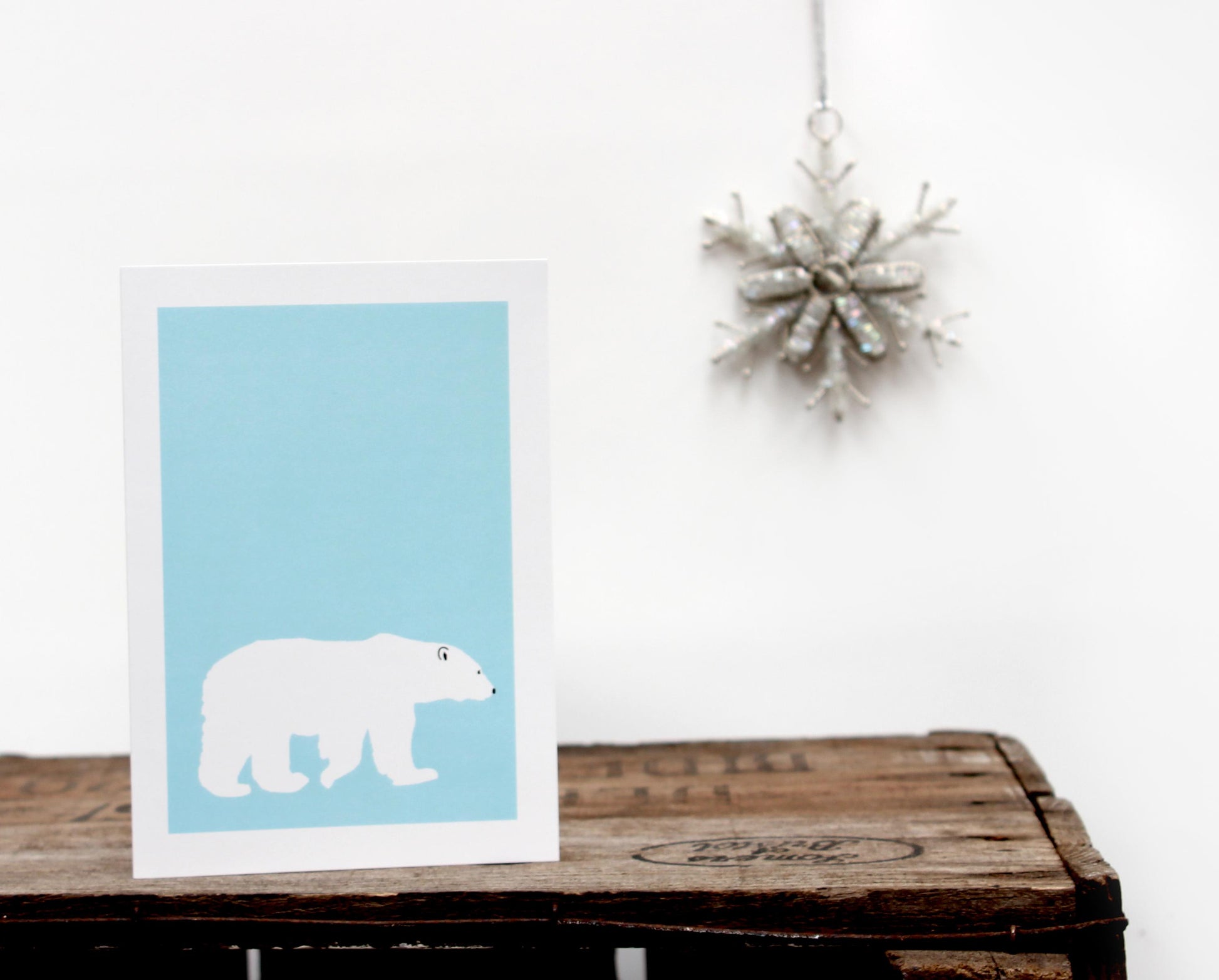 A greetings card with a blue background featuring a silhouette of a polar bear, placed on a wooden surface with a decorative Christmas tree in the background.