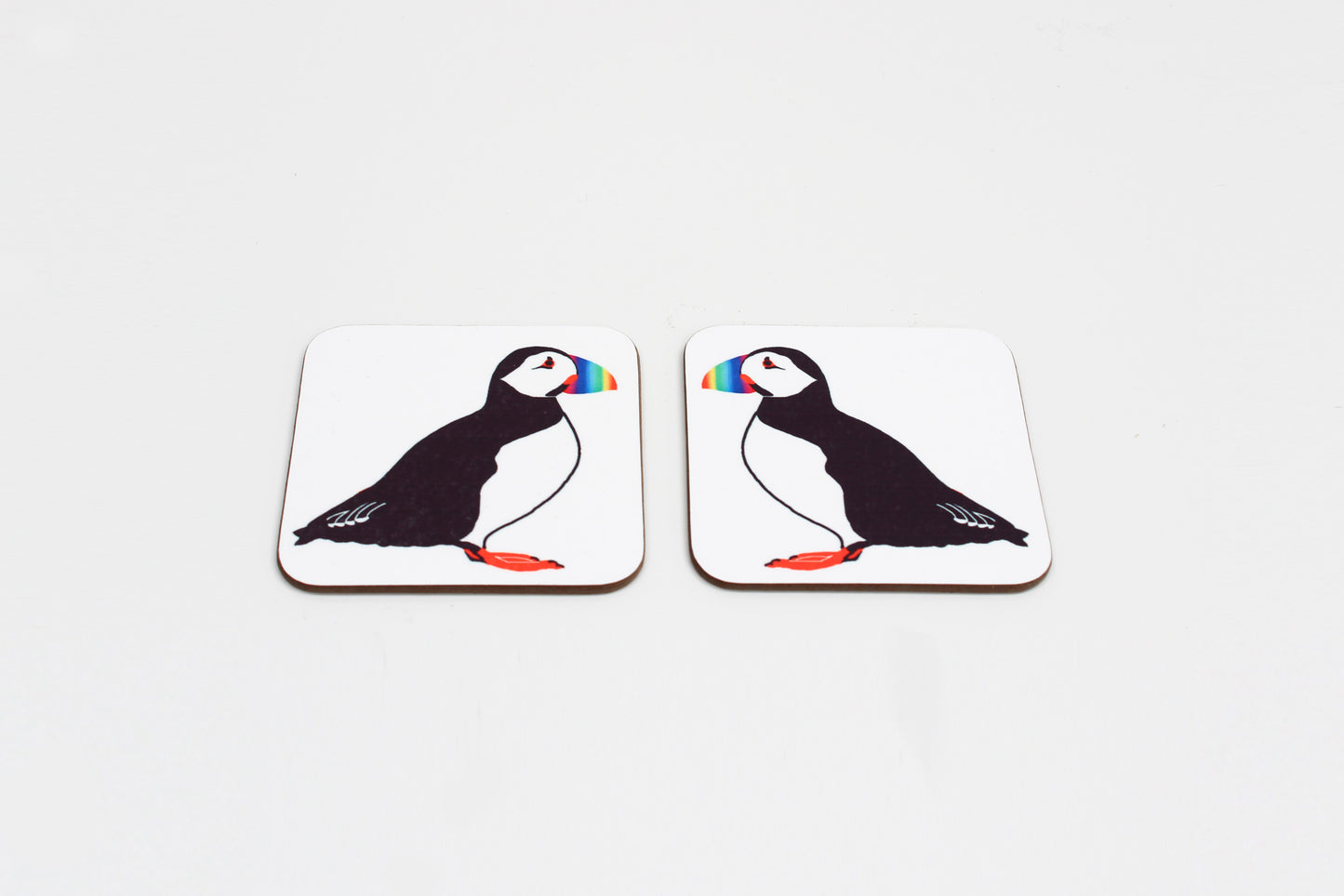 Two square coasters with a drawing of a puffin in the center with a rainbow-colored beak. Both have a white background.