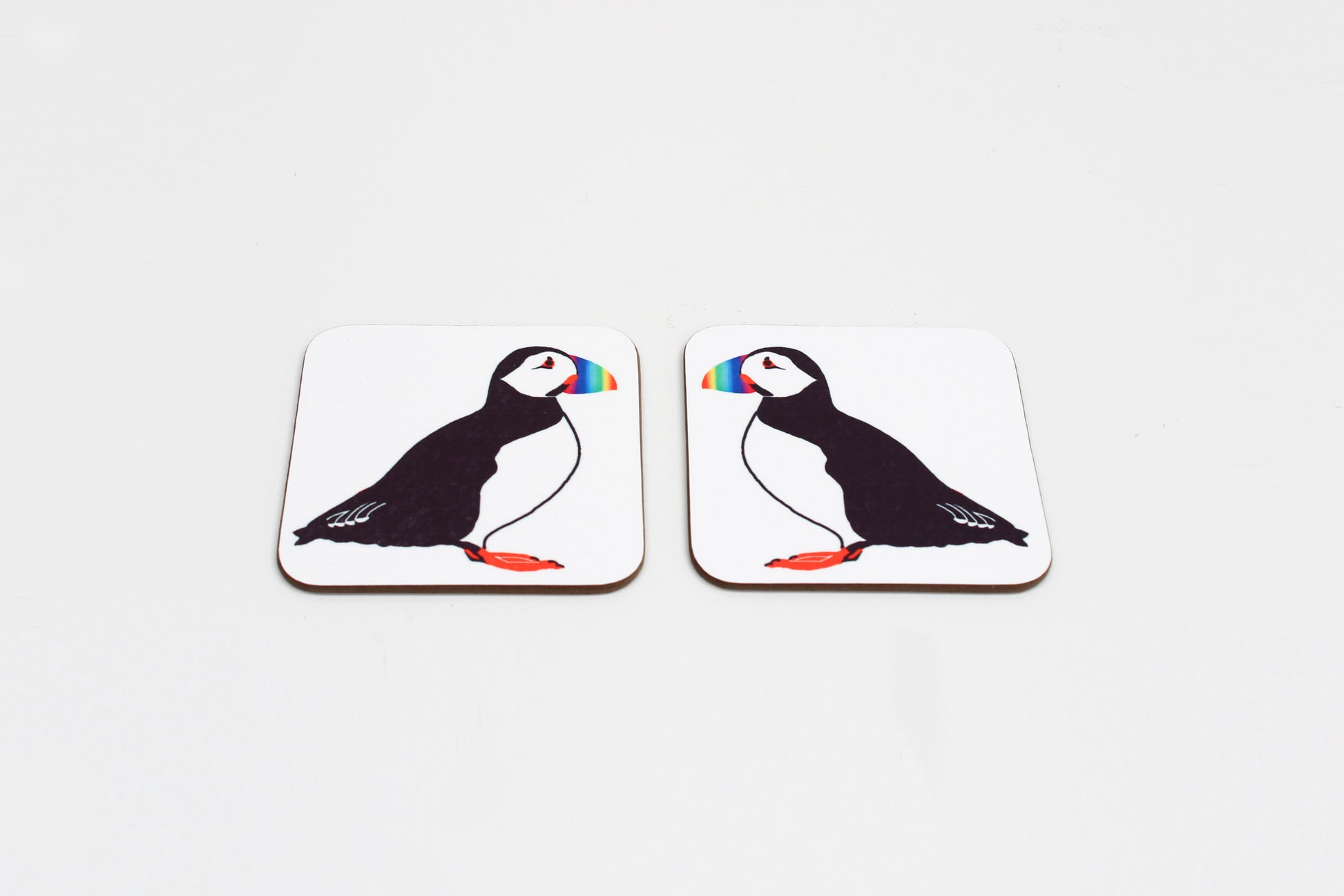 Two square coasters with a drawing of a puffin in the center with a rainbow-colored beak. Both have a white background.
