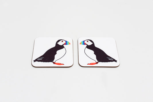 Two square coasters with a drawing of a puffin in the center with a rainbow-colored beak. Both have a white background.