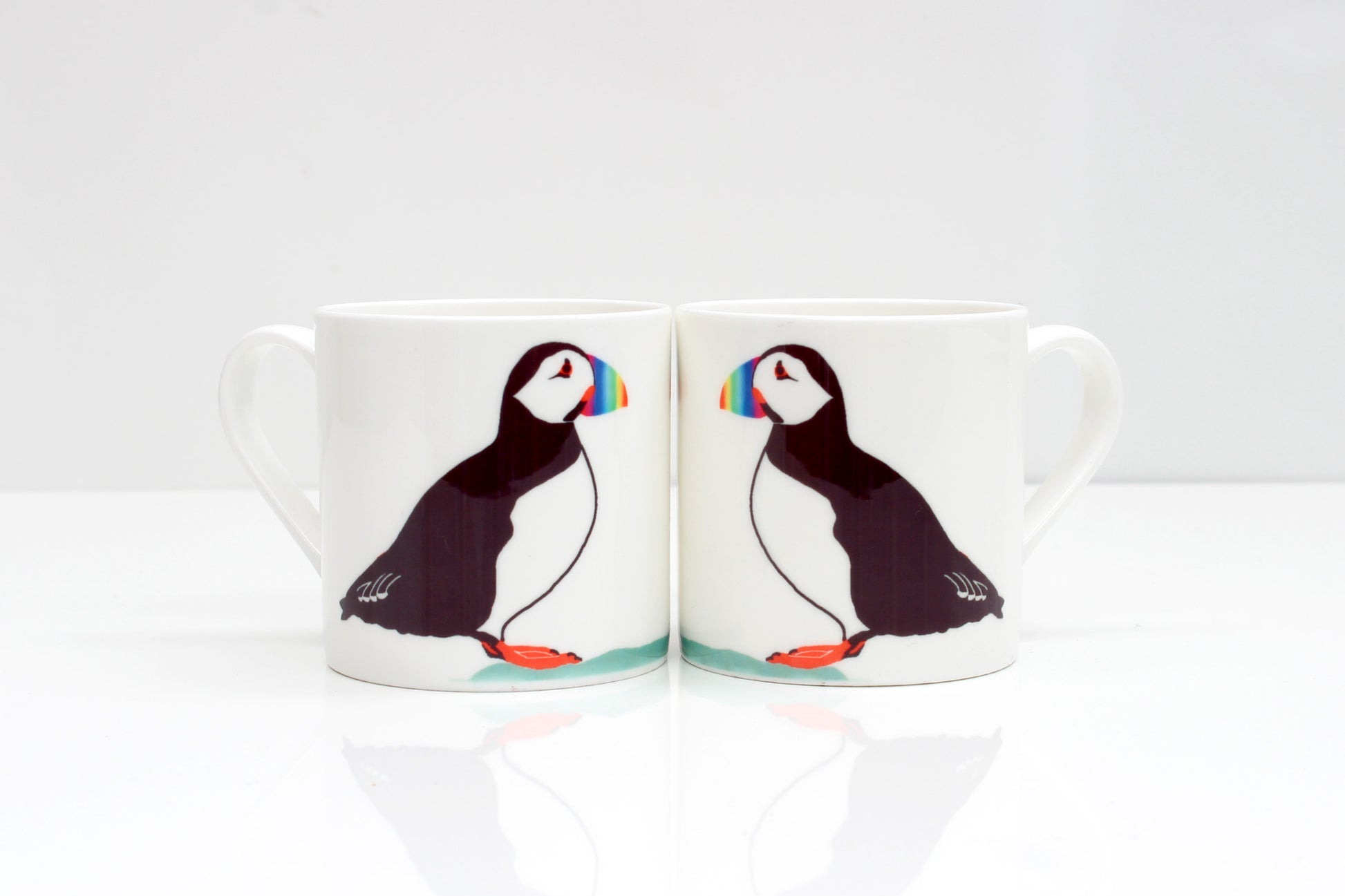 Two bone china mugs puffin designs, one mug showing the front side with the puffin, and the other mug showing the back side. Both mugs have a white color scheme and a reflective surface.