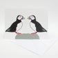 A greeting card with a illustration of two puffins facing each other. The card is set against a white background and there is a white envelope included.