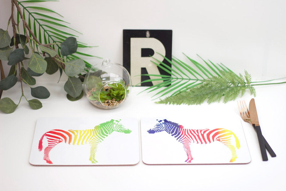 A set of placemats featuring a rainbow-colored zebra design, displayed on a table with a plant, a coaster, and a fork.