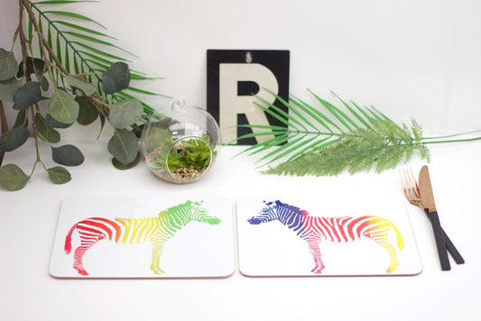 A set of placemats featuring a rainbow-colored zebra design, displayed on a table with a plant, a coaster, and a fork.