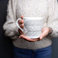 A person holding a white bone china mug with a geometric pattern.