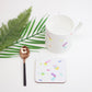 A white ceramic coaster with multicolored geometric patterns, a black spoon, and a piece of green foliage on a white background.