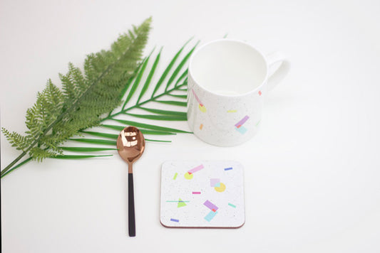 A white ceramic coaster with multicolored geometric patterns, a black spoon, and a piece of green foliage on a white background.