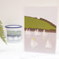 A greetings card featuring an illustration of sailboats on water, with a pastoral landscape in the background. The card is presented on a white surface with a small inset picture of a sailboat in a cup to the left.
