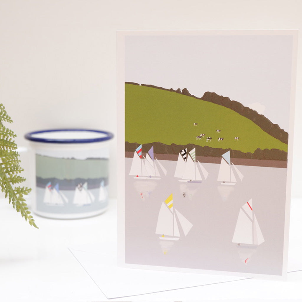 A greetings card featuring an illustration of sailboats on water, with a pastoral landscape in the background. The card is presented on a white surface with a small inset picture of a sailboat in a cup to the left.