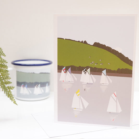 A greetings card featuring an illustration of sailboats on water, with a pastoral landscape in the background. The card is presented on a white surface with a small inset picture of a sailboat in a cup to the left.