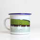 A white enamel mug with a blue rim, featuring a miss-printed design with boats and a pastoral landscape.