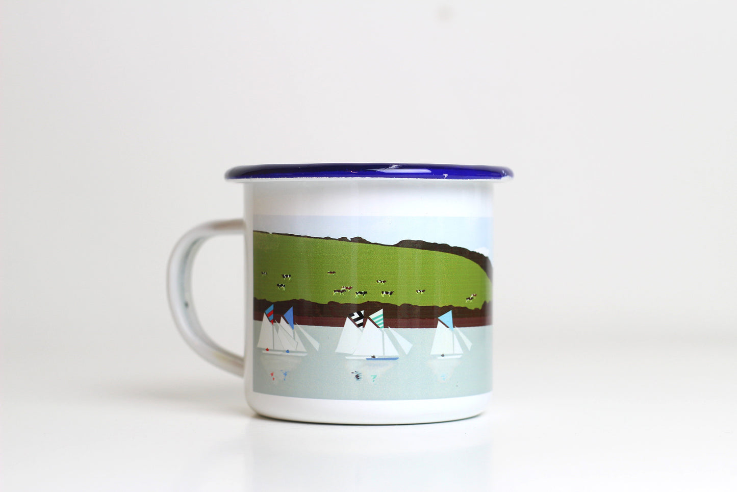 A white enamel mug with a blue rim, featuring a miss-printed design with boats and a pastoral landscape.