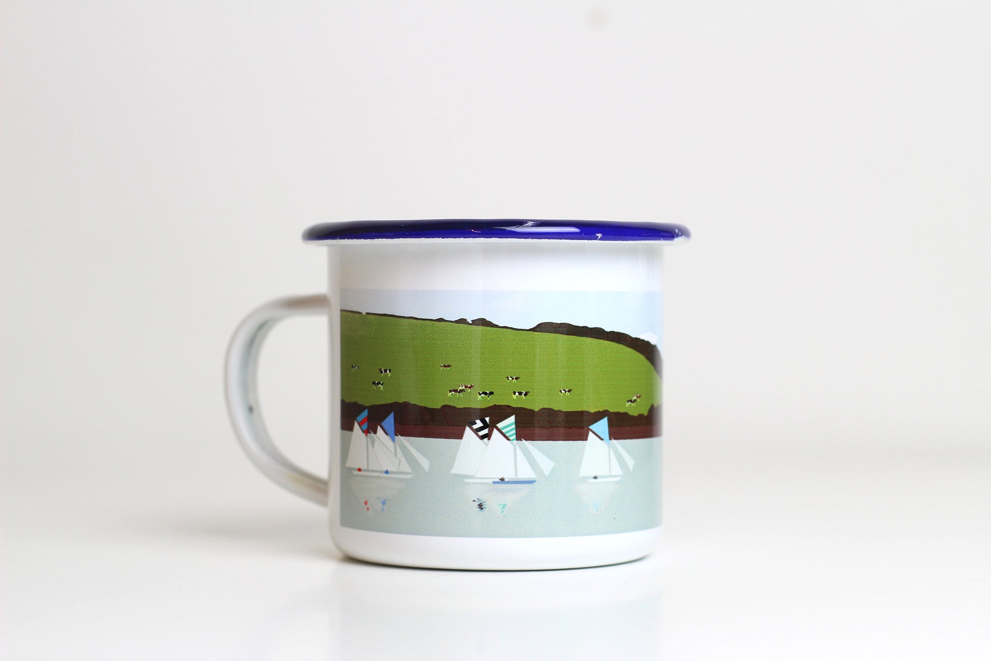 A white enamel mug with a blue rim, featuring a miss-printed design with boats and a pastoral landscape.
