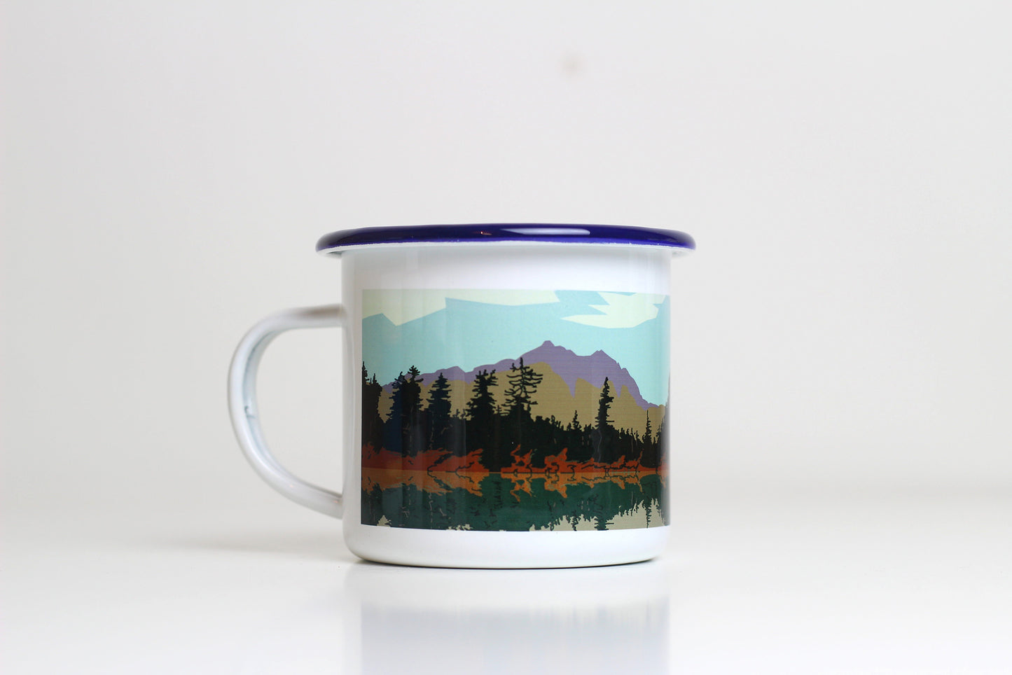 A white enamel mug with a blue lid, featuring a printed design with a mountain and pine forest landscape.