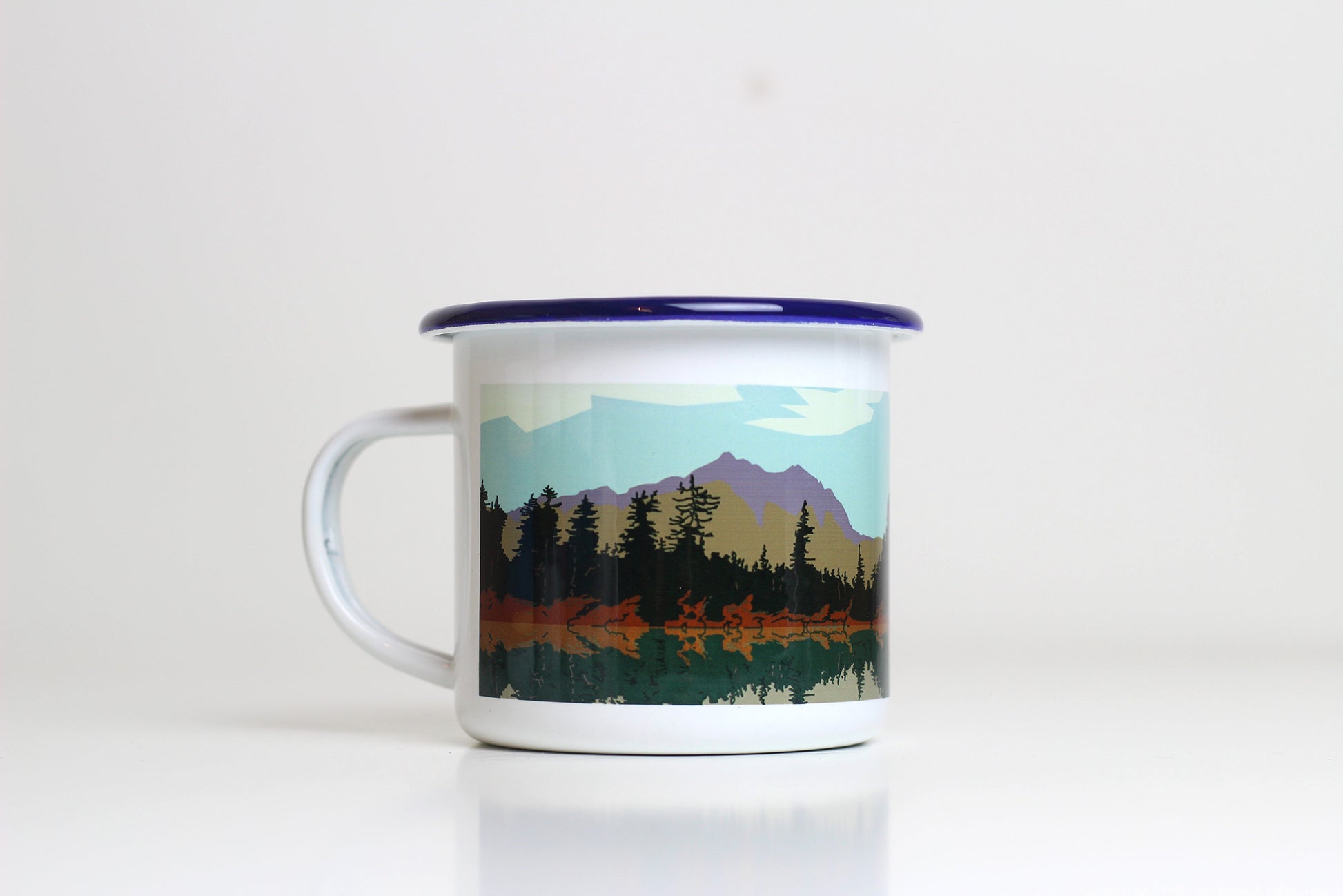 A white enamel mug with a blue lid, featuring a printed design with a mountain and pine forest landscape.