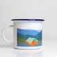 A white ceramic enamel mug with a blue rim, featuring a tent design and mountains in the image.