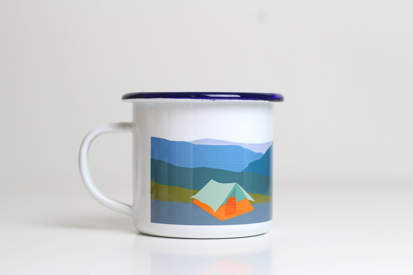 A white ceramic enamel mug with a blue rim, featuring a tent design and mountains in the image.