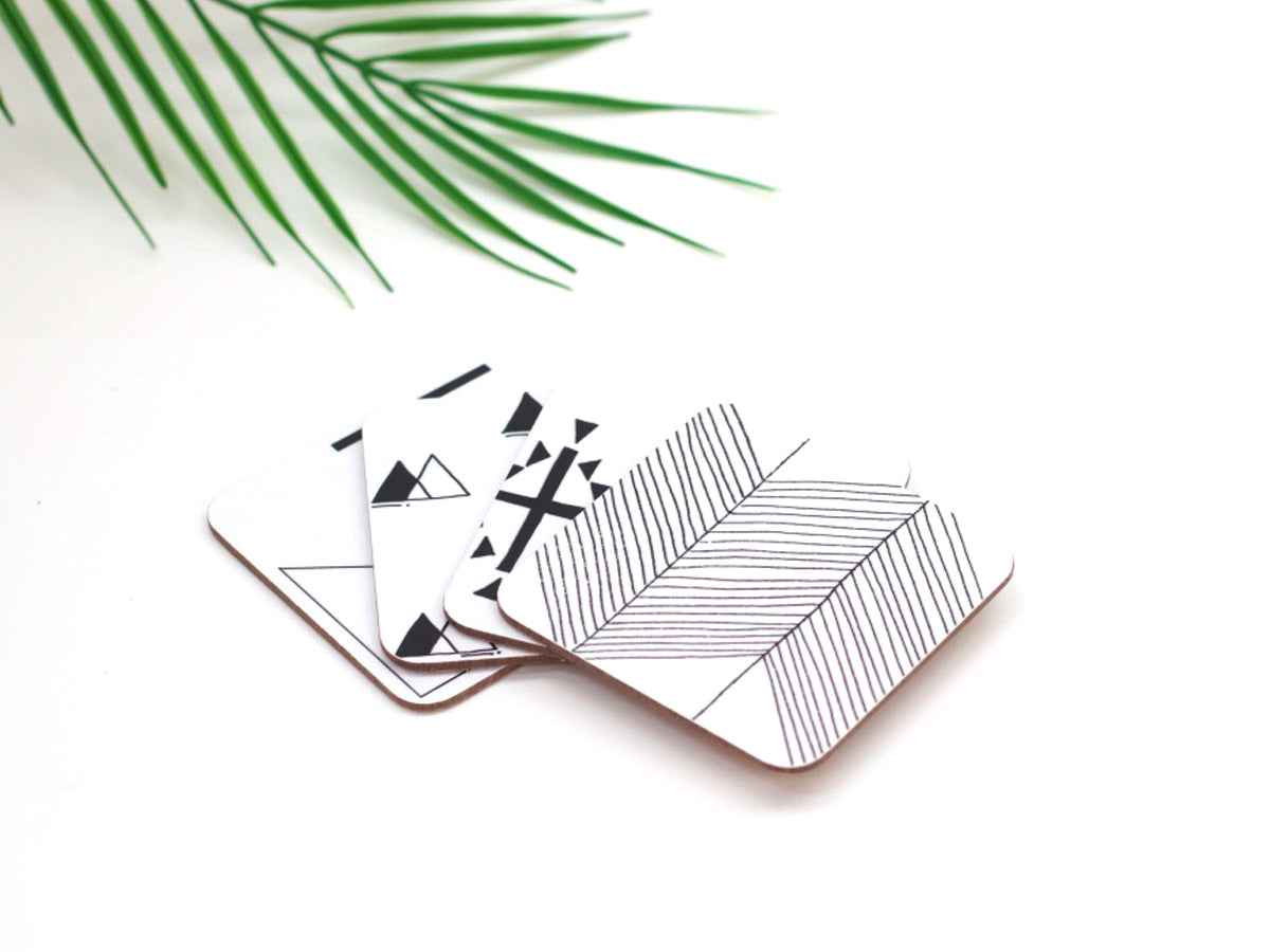A set of 4 square coasters with a geometric symbol design, placed on a white surface with a green leaves in the background.