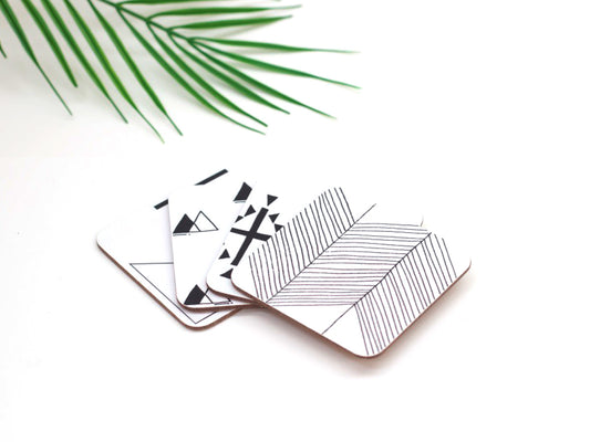 A set of 4 square coasters with a geometric symbol design, placed on a white surface with a green leaves in the background.