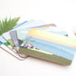 A collection of colorful placemats with various designs, including a beach scene and a forest, displayed on a table with a fork and a green plant in the background.
