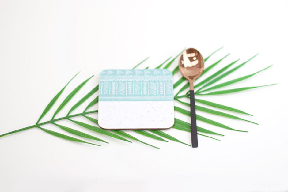 A blue and white geometric patterned coaster with a decorative element presented on a green palm leaf background.