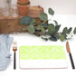 A rectangular placemat with a green and white geometric pattern, featuring a lime colour design, placed on a white surface with a fork and knife to the left, and a small potted plant and decorative leaves in the background.