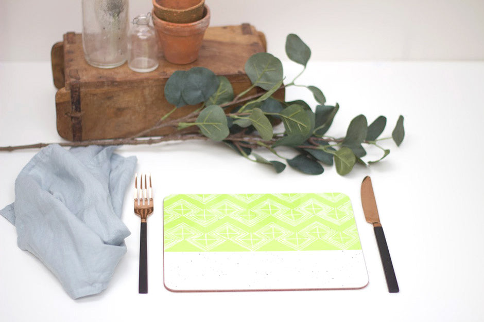 A rectangular placemat with a green and white geometric pattern, featuring a lime colour design, placed on a white surface with a fork and knife to the left, and a small potted plant and decorative leaves in the background.