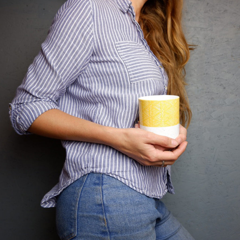 A person holding a white bone china mug with orange and yellow geometric patterns.