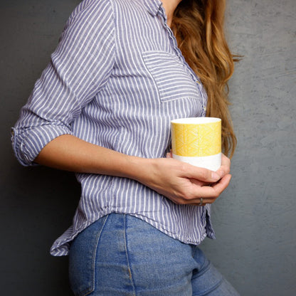 A person holding a white bone china mug with orange and yellow geometric patterns.