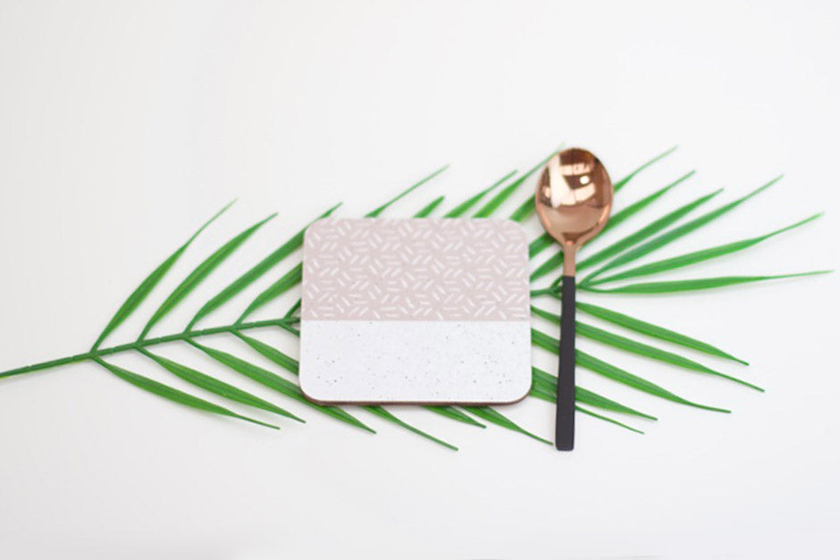 A square coaster with a geometric pattern, placed on a white surface alongside a spoon and a leaf.