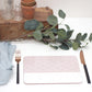 A rectangular placemat with a pink and white geometric pattern, a fork, knife, and a small napkin, displayed on a wooden board with a potted plant and decorative leaves in the background.