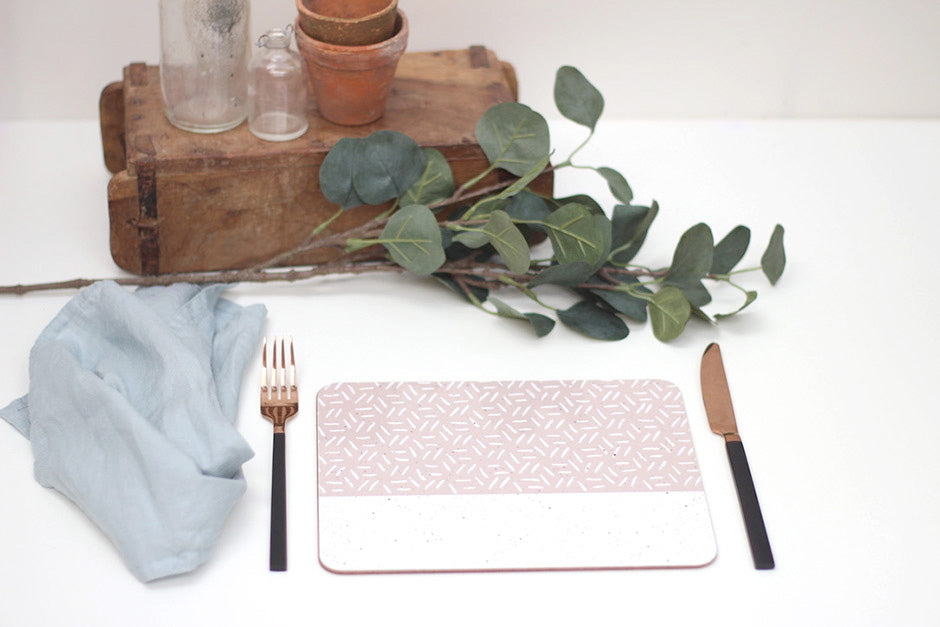 A rectangular placemat with a pink and white geometric pattern, a fork, knife, and a small napkin, displayed on a wooden board with a potted plant and decorative leaves in the background.