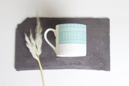 A bone china mug with a blue geometric pattern on a white background, accompanied by a dried plant on a slate background.