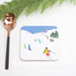 A coaster with a drawing of skiers on a snowy mountain, alongside a spoon, on a white background with green fern leaves in the background.