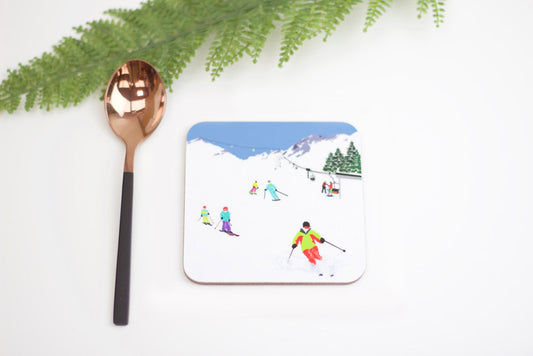 A coaster with a drawing of skiers on a snowy mountain, alongside a spoon, on a white background with green fern leaves in the background.
