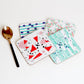 A set of four square coasters with various sound-inspired patterns including dots, lines, and geometric shapes, displayed on a white background alongside a teaspoon for scale.