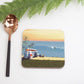 A square coaster with a beach and surfboard design, placed on a white surface next to a green fern leaf and a spoon.