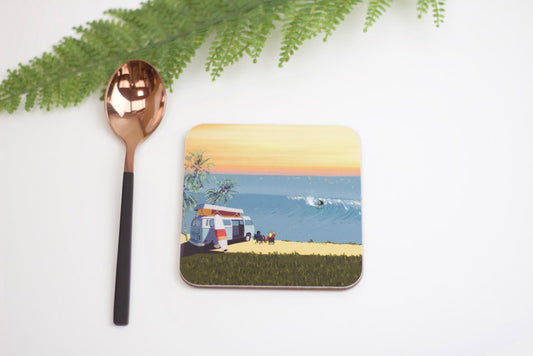 A square coaster with a beach and surfboard design, placed on a white surface next to a green fern leaf and a spoon.